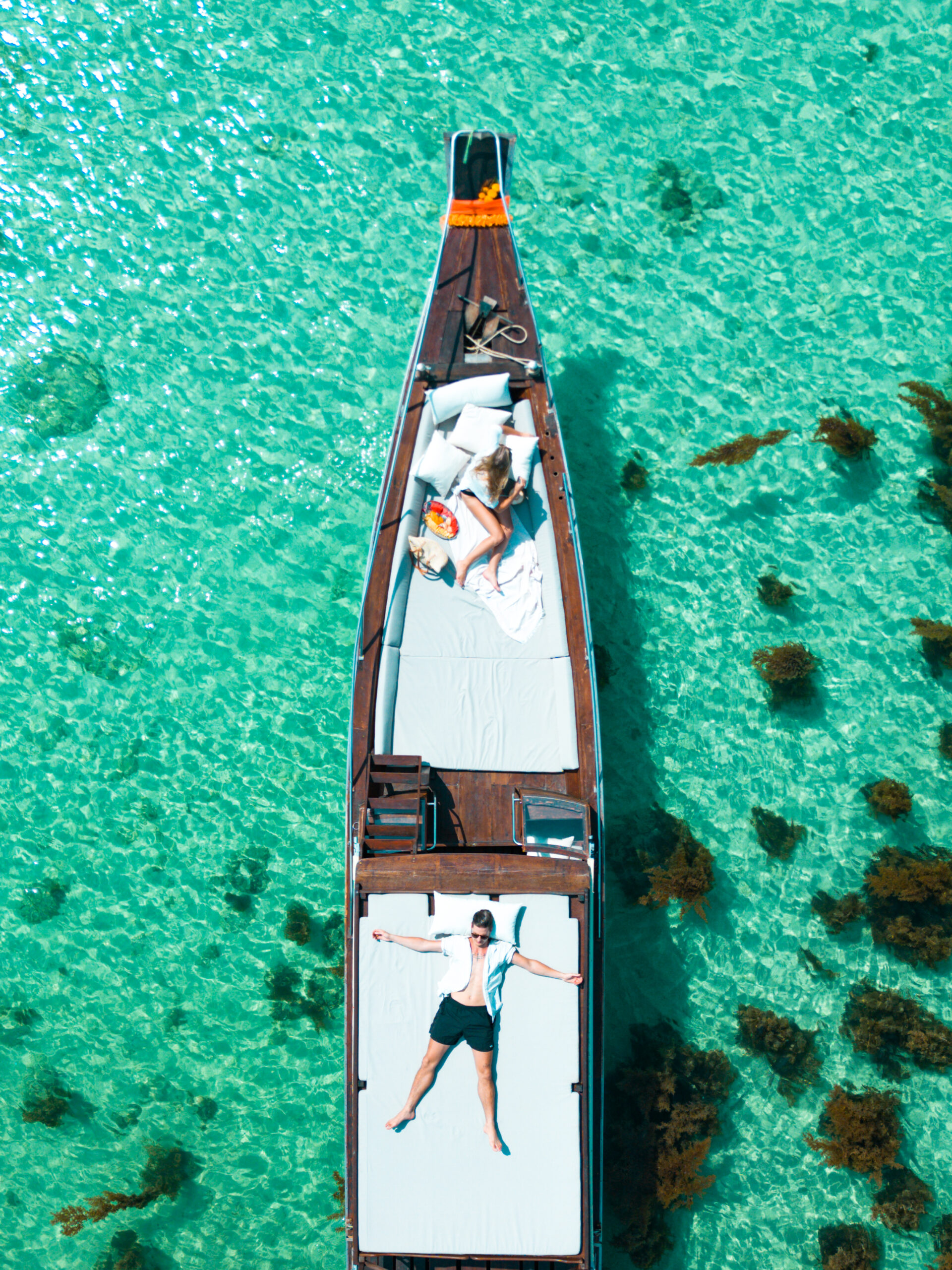 Aerial view of a long boat on clear turquoise waters with a person sunbathing and another person stretching on the deck.
