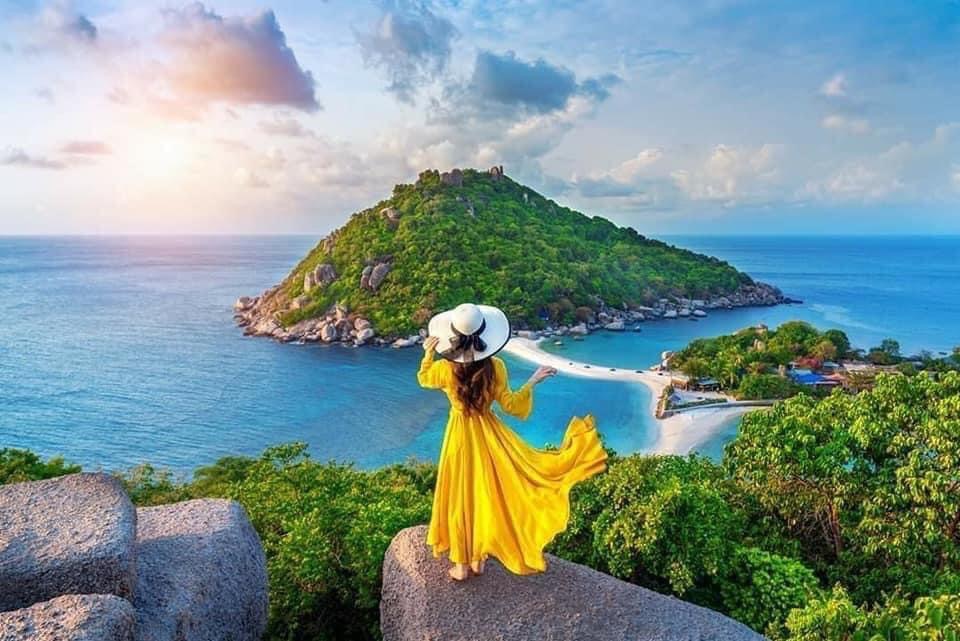 A woman in a flowing yellow dress and a white hat stands on a rocky overlook, gazing at the scenic view of a lush green island surrounded by blue sea.