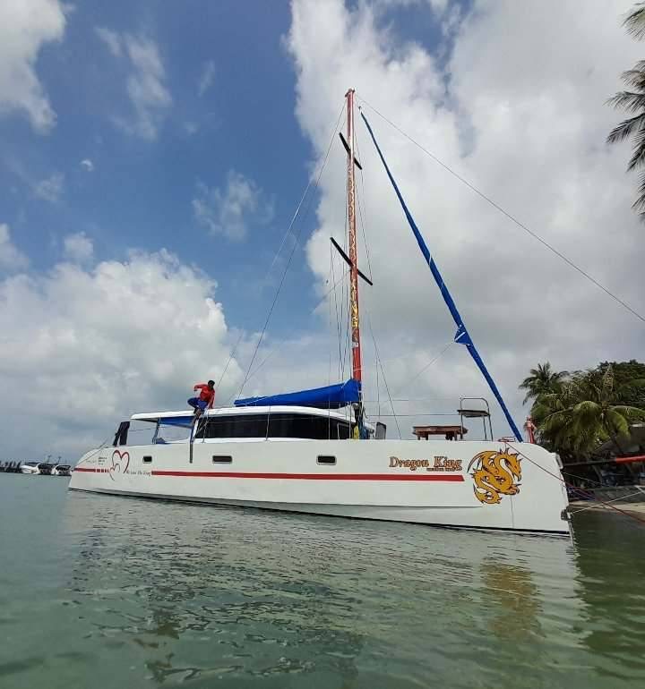 A white catamaran with red and blue details, named "dragon tales," floating on calm waters under a partly cloudy sky, with a person visible on its deck.