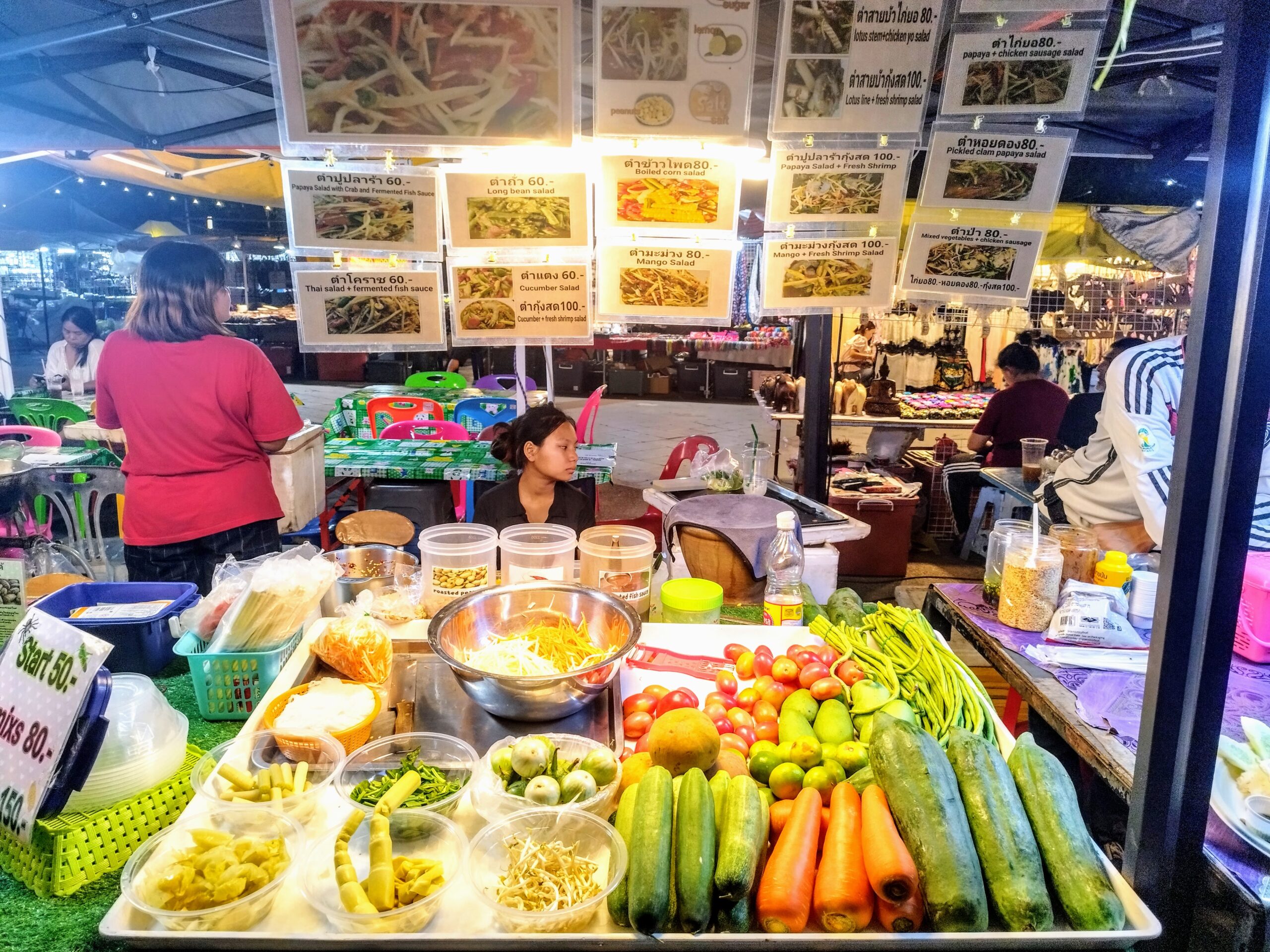 A market stall with various fresh vegetables, herbs, and containers of ingredients on a table. A woman sits behind the stall with menus displayed above. People are seen in the background.