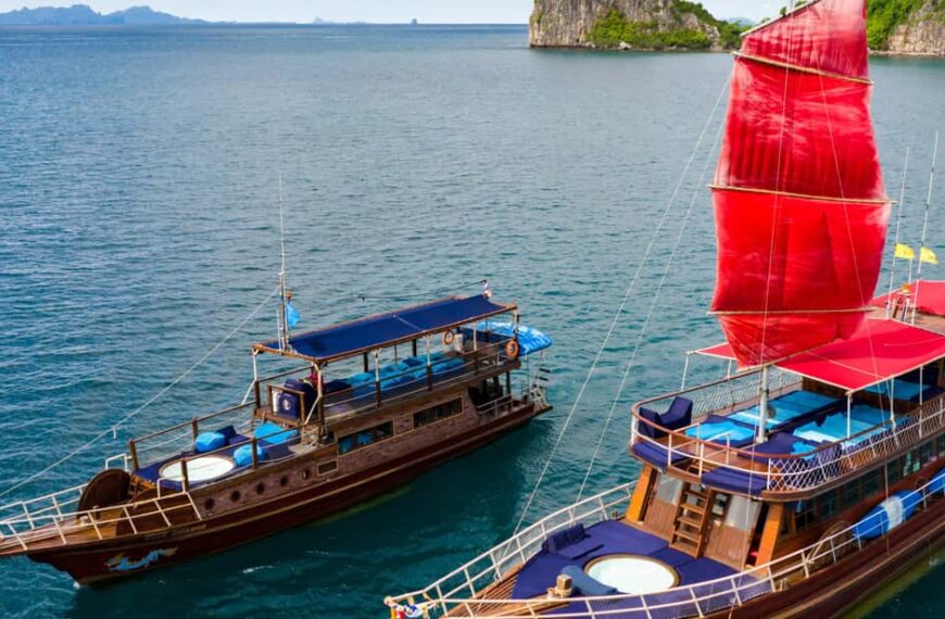 Two wooden boats with colorful awnings, including one with a red sail, are anchored on a calm sea with lush green cliffs in the background.