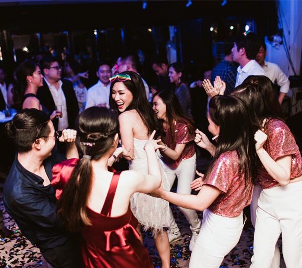 A group of people dance and socialize in a lively party setting. The room is dimly lit with blue light accents, and many attendees are smiling and enjoying the moment.