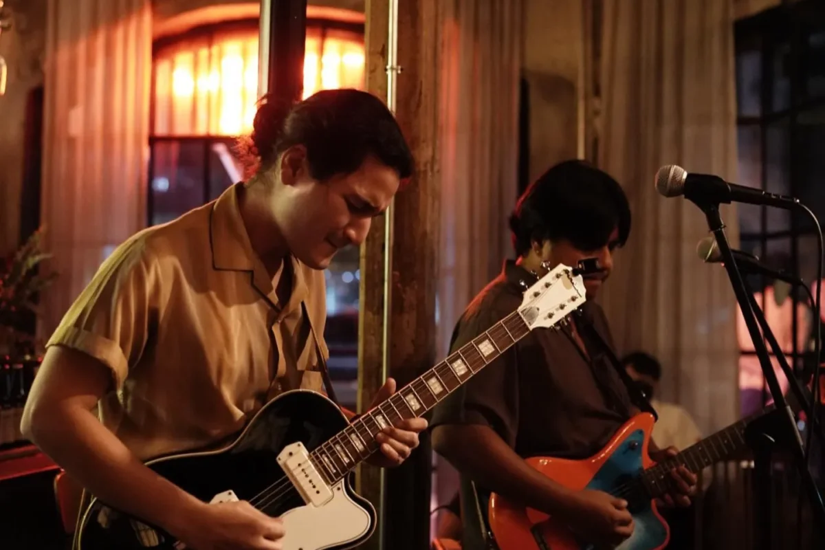 Two musicians are playing guitars in a dimly lit venue with a visible drum set in the foreground. One musician wears a beige shirt, while the other wears a darker shirt and sunglasses.