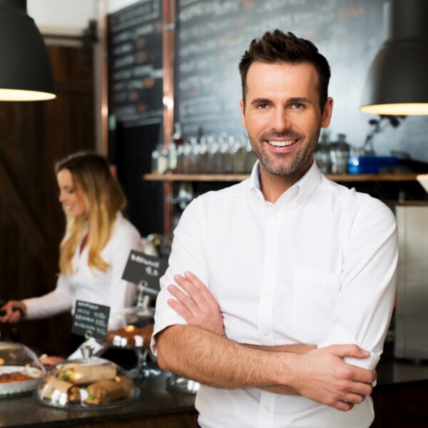 A man in a white shirt stands with arms crossed and smiles in a café; a woman is working at the counter in the background.