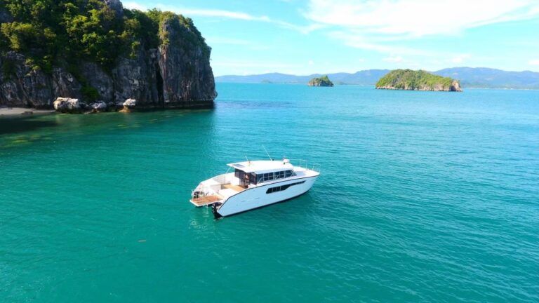 A white boat is anchored on clear turquoise water, near rocky cliffs and green islands under a blue sky.